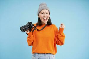 elegante asiático niña con digital cámara, tomando fotos. mujer fotógrafo sonriente, en pie terminado azul antecedentes foto