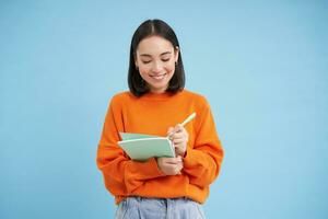 Smiling cute girl student, making notes, writing down homework, studying, standing over blue studio background photo