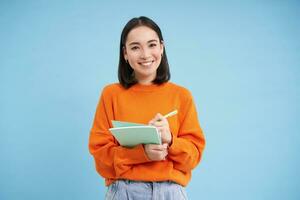 Beautiful asian girl, student taking notes, writing down, studying on courses, learning, standing over blue background photo