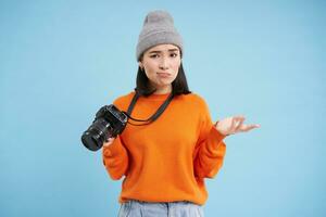 Portrait of asian woman in hat, holding digital camera with confused face, unprofessional photographer doesnt know how to take pictures on digicam, blue background photo