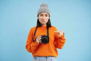 Asian girl in hat, points at her digital camera with disappointed, upset face, doesn't like her device, photographer complains at her digicam, blue background photo