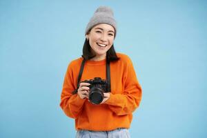 Stylish asian girl with digital camera, taking pictures. Woman photographer smiling, standing over blue background photo