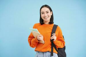 educación y estudiantes. sonriente joven asiático mujer con mochila y cuadernos, posando en contra azul antecedentes foto