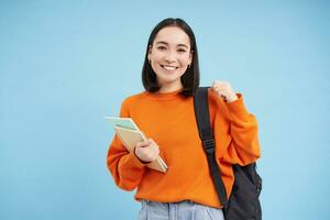 educación y estudiantes. sonriente joven asiático mujer con mochila y cuadernos, posando en contra azul antecedentes foto