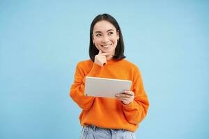 Woman with digital tablet thinking, looking aside with thoughtful face expression, standing over blue background photo