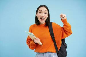 Enthusiastic girl student, celebrating, wearing backpack and holding notebooks, college homework, standing over blue background photo