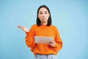 Portrait of unsure girl, asian woman with digital tablet, shrugging shoulders and looks complicated, standing over blue background photo