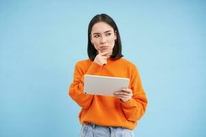 Woman with digital tablet thinking, looking aside with thoughtful face expression, standing over blue background photo