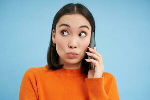 Technology concept. Cute korean woman talks on mobile phone, looks aside with surprised face, stands over blue background photo