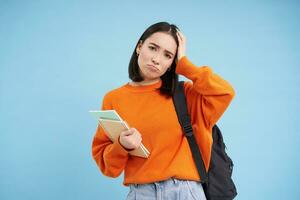 Sad girl student, touches her head and sulks, looks tired after college, holds backpack and notebooks, blue background photo