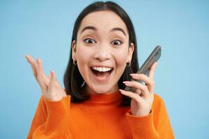 Girls reaction to amazing news she received on mobile phone. Happy asian woman looks surprised and excited, blue background photo