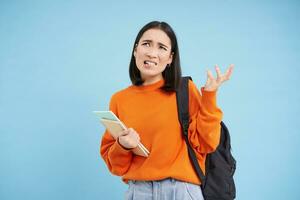Annoyed young asian woman, student complans, shakes hand and looks disappointed, stands with backpack and notebooks, blue background photo
