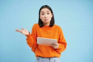 Portrait of unsure girl, asian woman with digital tablet, shrugging shoulders and looks complicated, standing over blue background photo