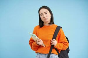 Sad sulking asian student, girl with backpack and notebooks, looking upset, standing over blue background photo