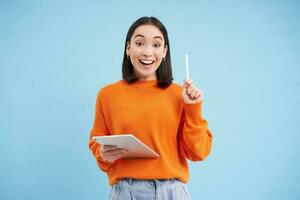 Eureka, got an idea. Excited asian woman with digital tablet, raising pen up, got revelation, has great solution, stands over blue background photo