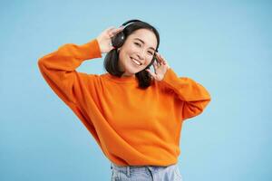 hermosa coreano mujer en auriculares, bailando y escuchando música en auriculares, en pie terminado azul antecedentes foto