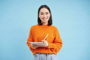 Portrait of young Chinese woman, teacher or student with digital tablet and pencil, writing, taking notes, doing her homework, standing over blue background photo