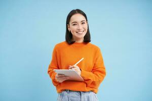 Portrait of young Chinese woman, teacher or student with digital tablet and pencil, writing, taking notes, doing her homework, standing over blue background photo