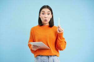 Eureka, got an idea. Excited asian woman with digital tablet, raising pen up, got revelation, has great solution, stands over blue background photo