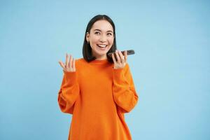 alegre sonriente coreano mujer hablar en altavoz, registros voz mensaje, aprende cómo a hablar utilizando idioma aprendizaje móvil aplicación, azul antecedentes foto