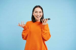 Happy asian woman records voice message, repeats words in language learning mobile app, talks in smartphone speakerphone, blue background photo
