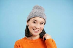 cerca arriba retrato de sonriente asiático mujer en calentar sombrero, mirando contento y linda a cámara, tiene claro natural piel, soportes terminado azul antecedentes foto