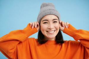 Close up portrait of happy, stylish korean girl puts on warm hat, smiles and looks joyful, stands against blue studio background photo