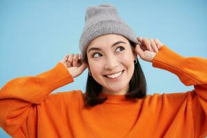 Close up portrait of smiling asian woman in warm hat, looking happy and cute at camera, has clear natural skin, stands over blue background photo