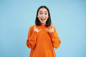 Portrait of excited young woman, shows rock n roll, heavy metal party sign, enjoys event, having fun, standing over blue background photo