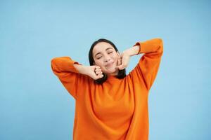 Beautiful asian girl, 25 years, stretches her arms with pleased lazy face, wakes up after sleep, good nap, stands over blue background photo