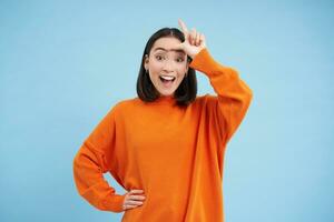 Enthusiastic korean girl, shows L letter on forehead and laughs, mocks someone who lost, stands over blue background photo