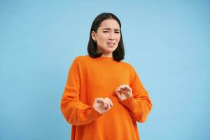 Disgusting. Asian korean girl cringes, grimaces from aversion and dislike, blocks her hands, steps back from disgust, stands over blue background photo