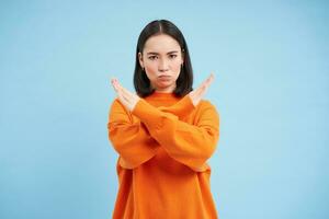 imagen de grave coreano mujer muestra cruzar manos gesto, detener firmar, tabú, desaprueba malo acción, soportes en naranja camisa terminado azul antecedentes foto