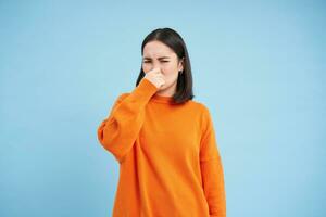 Something stinks. Young asian woman shuts her nose with fingers from bad smell, wears orange sweatshirt, stands over blue background photo