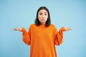 I dont know. Puzzled asian woman shrugs shoulders with hands spread sideways, confused, dont know what to do, clueless and unaware what happened, blue background photo