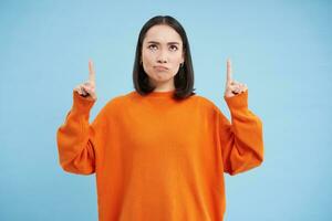 Perplexed asian girl, looks and points up with frustrated, thinking face expression, shows advertisement and reads it with doubt, stands over blue background photo