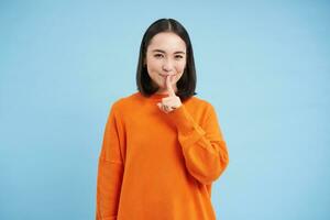 Hush its a secret. Young asian woman shushing, asks to be quiet, holds finger on lips, gossips, stands in orange sweatshirt over blue background photo