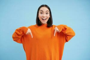 Cheerful asian woman shows advertisement below, points down with smiling face, inviting people to click on link, follow arrow, standing over blue background photo