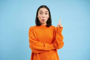 Eureka, I got an idea. Excited korean woman shows one finger pointing up, looks amazed, has plan or revelation, says breaking news, stands over blue background photo