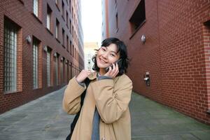 retrato de joven coreano mujer caminando abajo calle con teléfono, hablando con alguien, hace un llamar, tiene teléfono conversacion foto