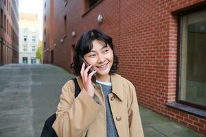 retrato de joven coreano mujer caminando abajo calle con teléfono, hablando con alguien, hace un llamar, tiene teléfono conversacion foto