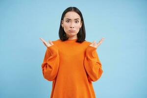 Asian girl with confused face, shrugs shoulders and pouts, looks upset and clueless, dont understand, puzzled by smth, stands over blue background photo