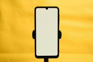 Portrait phone with white screen fixed to tripod on yellow background, for mockup design. photo