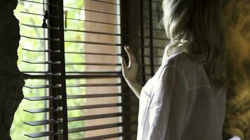 Charming young blonde woman touching the blinds by her hand, looking out the glass window. Art. Young model looking pensive and standing by the window. photo