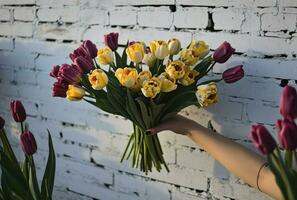 ai generado persona participación ramo de flores de amarillo y púrpura tulipanes en blanco ladrillo foto