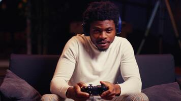 BIPOC man in living room playing videogame on gaming console, spamming controller buttons. Gamer wearing headphones to communicate with teammates while battling enemies in online multiplayer shooter video