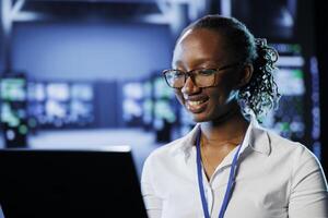 Joyful admin in high tech workplace uses laptop to prevent system overload. Competent expert between server racks ensuring enough network bandwidth for smooth operations photo