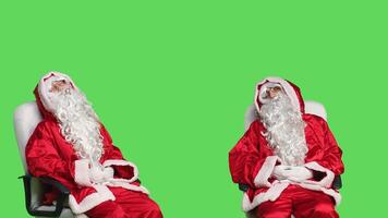 Father christmas falling asleep on chair, trying to keep eyes open in studio with greenscreen. Man in saint nick costume sleeping on camera, feeling tired and relaxing before festive celebration. photo