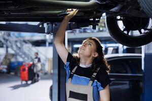 meticuloso mecánico trabajando en suspendido coche en cochera, comprobación motor durante rutina mantenimiento. auto reparar tienda empleado debajo vehículo, inspeccionando partes utilizando trabajo ligero foto