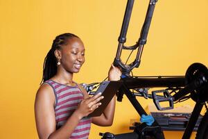 joven negro dama inspeccionando dañado bicicleta y explorador reparar opciones en su inteligente tableta. imagen de africano americano mujer comprobación roto bicicleta componentes mientras participación un digital dispositivo. foto